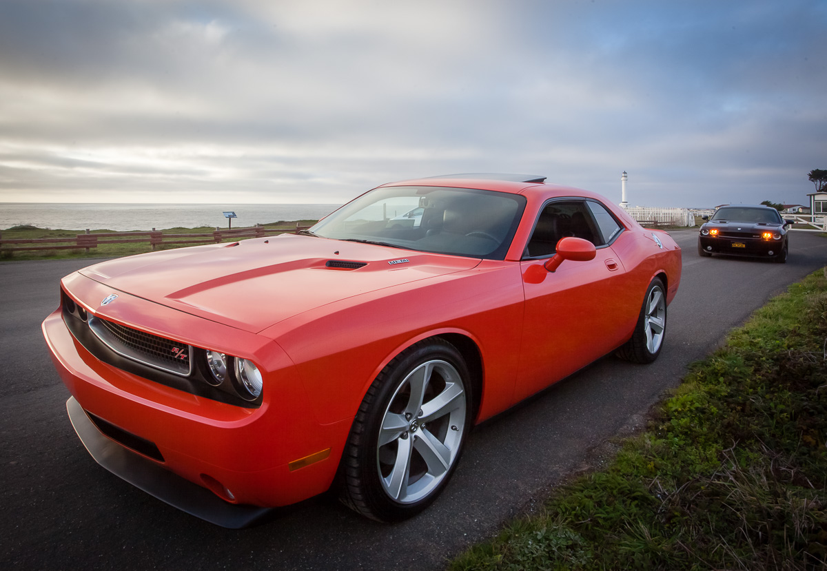 Red Dodge Challenger muscle car parked on a coastal road with a black Dodge Challenger behind it, ocean and cloudy sky in the background.