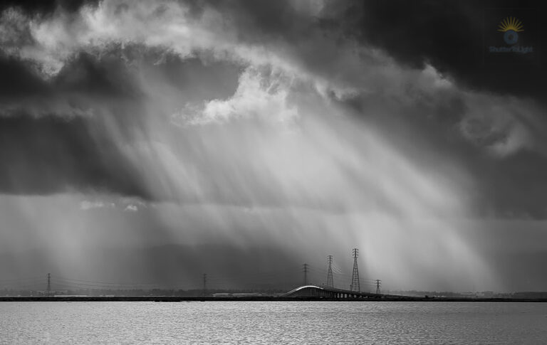 Beams of sunlight slice through stormy clouds, lingering over the bridge and power towers—a fleeting moment of stillness and anticipation.