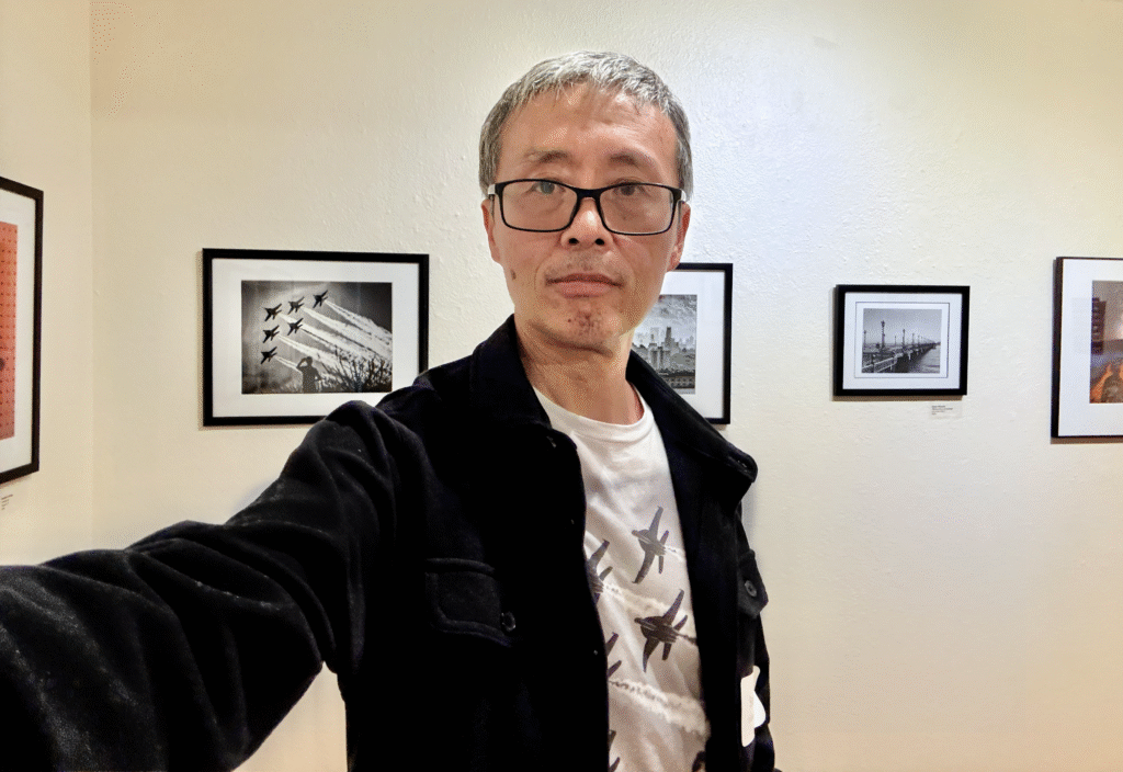 Photographer standing in front of framed black-and-white aviation photograph at Olive-Hyde exhibition, wearing a jacket and printed T-shirt matching the photo.