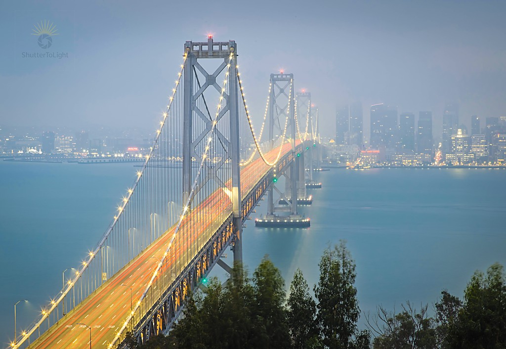 Long‑exposure view of the Bay Bridge at dusk, with continuous red tail‑light trails running across the span, dark trees in the foreground, and the distant city skyline fading into blue‑gray mist above calm water.