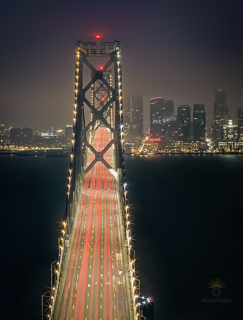 Nighttime long‑exposure photograph looking straight down the Bay Bridge toward a distant city skyline, with red car tail‑light trails forming bright lines along the roadway, bridge towers outlined by rows of star‑shaped lights, and the surrounding bay fading into dark blue haze.