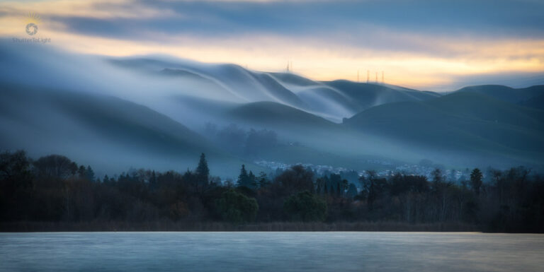 Fog lifting from Fremont's Lake Elizabeth at dawn: smooth water, layered mist over hills, warm sky glow above cool haze. Long exposure captures atmospheric transition.