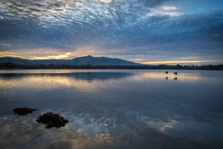 Early morning lake with calm water reflecting a fading red sunrise, blue clouds overhead, and distant birds silhouetted against the bright horizon, creating a quiet, still atmosphere.