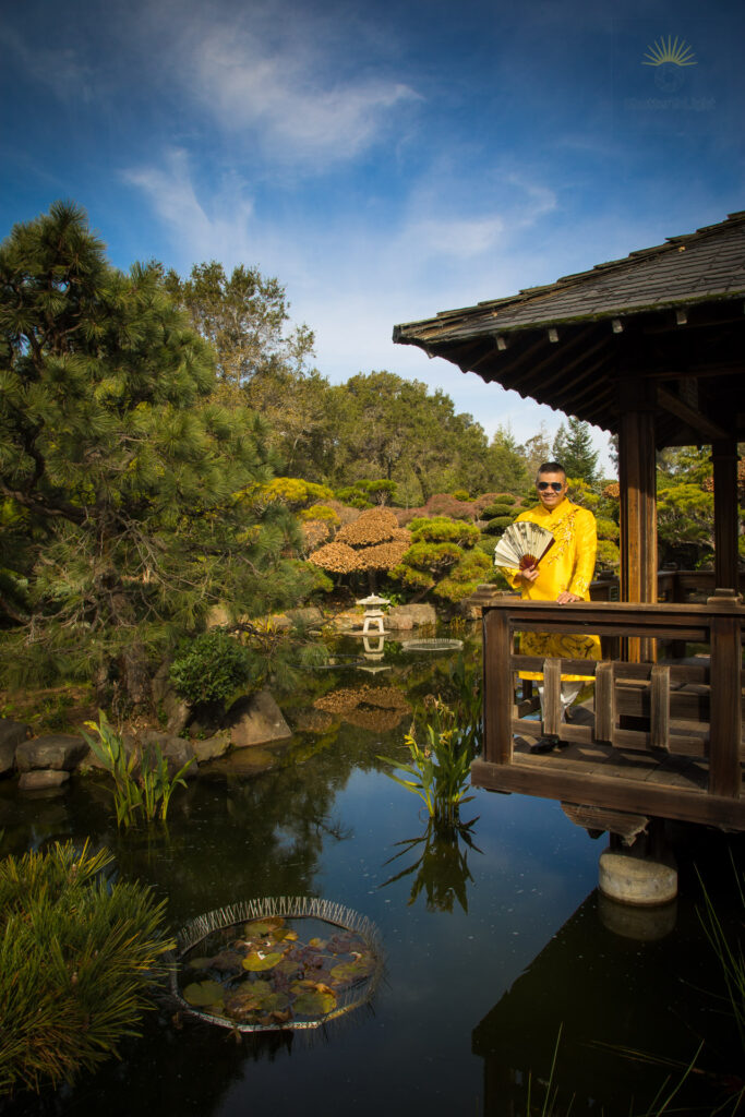 Uy Dinh wearing a gold traditional Vietnamese áo dài stands on a wooden pavilion overlooking a reflective pond at the Hayward Japanese Garden, surrounded by manicured trees and calm water.