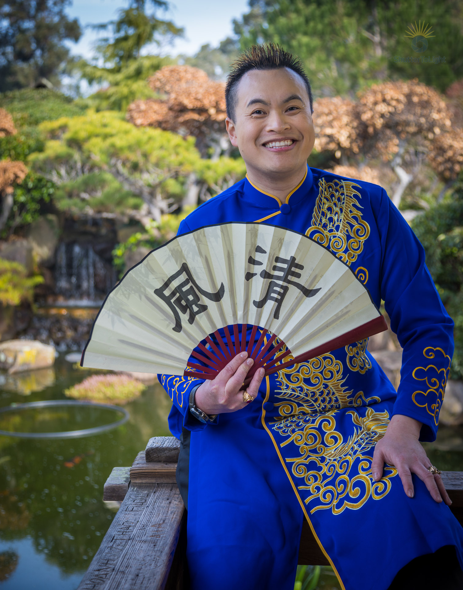 Trung Bui in a deep blue Vietnamese áo dài with golden dragon embroidery, holding a fan inscribed with 清风 meaning 'pure wind,' photographed near the pond surrounded by layered greens and waterfall.