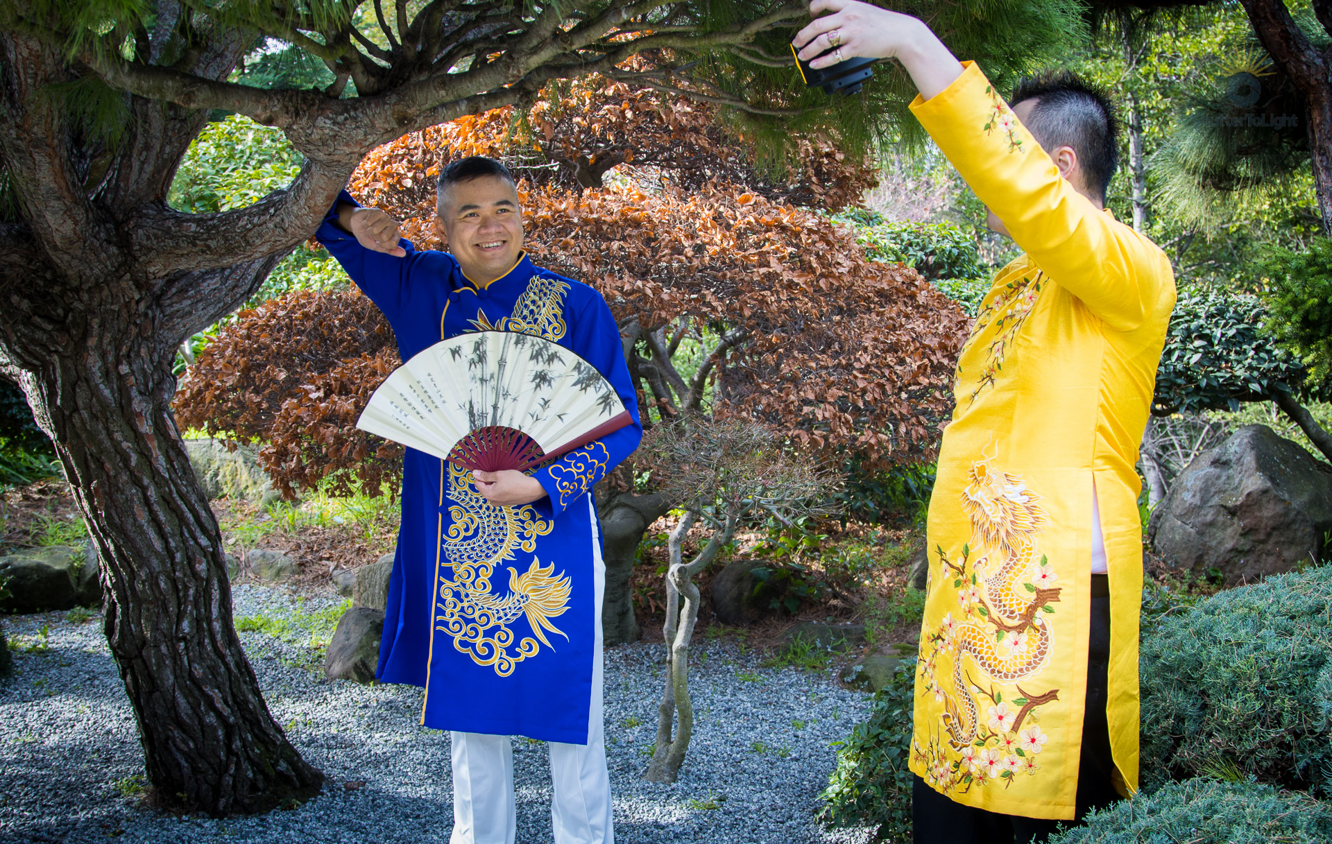 Uy Dinh in royal blue áo dài with gold dragon motif holds an ornate fan as Trung Bui in bright yellow áo dài lifts an LCD light panel to illuminate her, beneath a textured tree in lush Hayward Japanese Garden greenery.