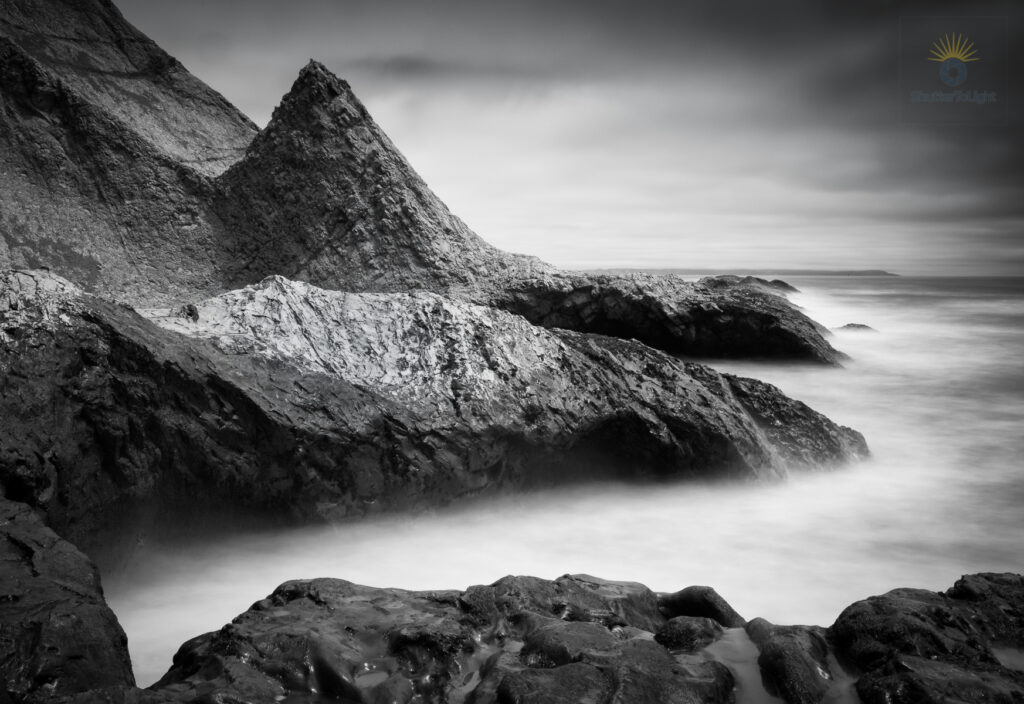 Long‑exposure black‑and‑white photograph of a triangular sea stack rising from the ocean at Agate Beach, with misty water created by a 30‑second exposure surrounding the sharp rock under soft overcast light.