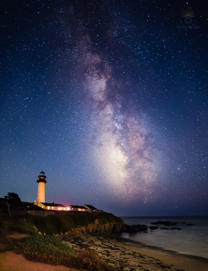 The Milky Way rises above Pigeon Point Lighthouse, with a beach, rocky coastline, and ocean beneath the night sky.